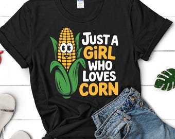 May include: Black t-shirt with the text "Just a Girl Who Loves Corn" and a cartoon corn graphic. The shirt is paired with denim shorts and sandals, creating a casual, everyday look.
