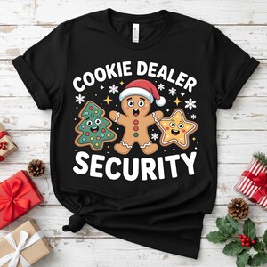 May include: Black t-shirt with the text "COOKIE DEALER SECURITY" and cartoon gingerbread man, Christmas tree, and star designs. The gingerbread man wears a Santa hat. Festive holiday apparel.