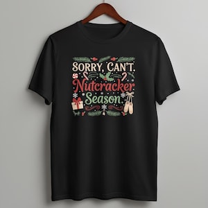 May include: Black t-shirt with the text "SORRY, CAN'T. Nutcracker Season." The design includes candy canes, holly, a gift, snowflakes, and ballet shoes. The shirt is hanging on a wooden hanger.