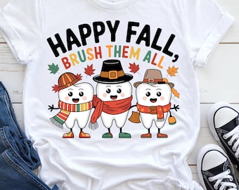 May include: White t-shirt with a fall-themed graphic. The design features three cartoon teeth wearing hats and scarves, with the text "HAPPY FALL, BRUSH THEM ALL" in black and orange. Autumn leaves surround the text and characters.