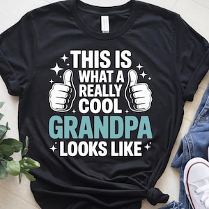 以下が含まれることがあります： 白い文字で「This is what a really cool Grandpa looks like.」と書かれた黒いTシャツ。  テキストの両側に白い親指が2本あります。  テキストの周りに小さな白い星があります。