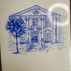 May include: A white ceramic tile with a blue ink drawing of a classical building. The building has columns, windows, and a detailed facade. A tree is drawn on the left side of the building.