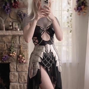 May include: A black sleeveless dress featuring a metallic chainmail shoulder piece, a cutout bodice with crisscross straps, and a layered sheer skirt. The outfit includes a silver-toned segmented waist armour piece with hanging chain details.