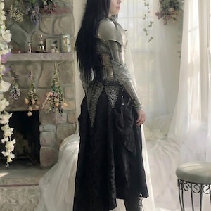 May include: A person wearing silver armor over a black dress. The armor covers the shoulders and arms. The black dress has a long skirt. The person is standing near a window with white curtains.