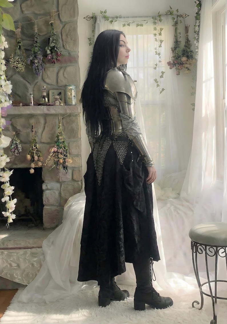 May include: A person wearing silver armor over a black dress. The armor covers the shoulders and arms. The black dress has a long skirt. The person is standing near a window with white curtains.