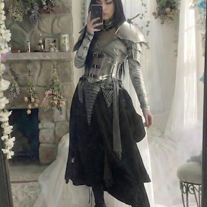 May include: A silver and black medieval-inspired outfit. The ensemble includes a metallic breastplate, shoulder armor, and a long black skirt. The person is holding a phone up to take a selfie in a mirror.