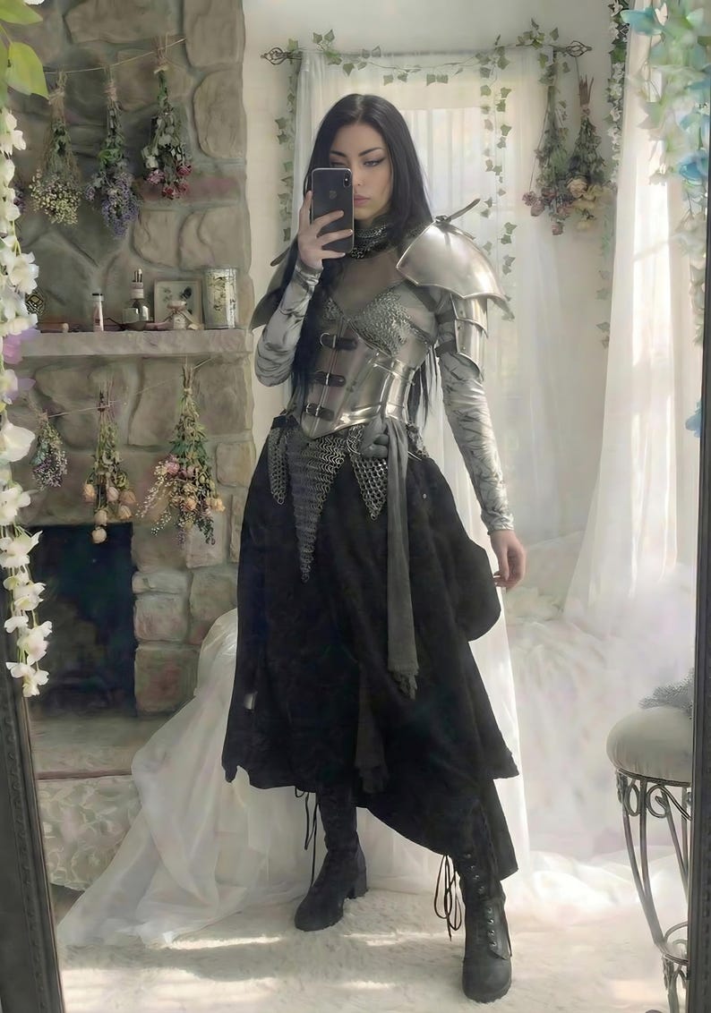 May include: A silver and black medieval-inspired outfit. The ensemble includes a metallic breastplate, shoulder armor, and a long black skirt. The person is holding a phone up to take a selfie in a mirror.