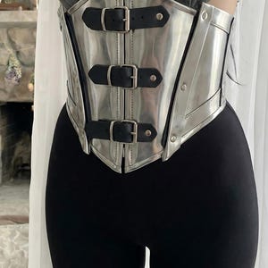 May include: A silver-colored corset with three black leather straps and buckles. The corset has a shiny, metallic finish and is worn over a black top and leggings. The design is reminiscent of armor.