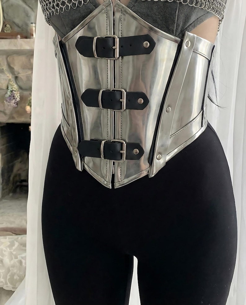 May include: A silver-colored corset with three black leather straps and buckles. The corset has a shiny, metallic finish and is worn over a black top and leggings. The design is reminiscent of armor.