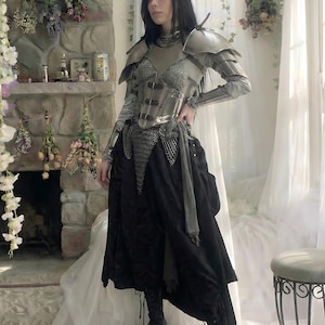 May include: A silver and black medieval-inspired outfit. The ensemble includes a metallic breastplate, shoulder armor, and chainmail accents. A black skirt and boots complete the look. The outfit is designed with a fantasy theme.