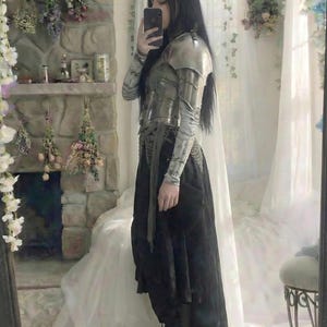 May include: A person wearing a silver-toned metal armor breastplate and shoulder guards over a long-sleeved shirt, paired with a black skirt and boots. The outfit is reflected in a mirror, with a stone fireplace and floral decorations in the background.