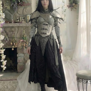 May include: A person wearing a silver-toned medieval-style armor set, including a breastplate, shoulder guards, and chainmail accents. The outfit is paired with a black skirt and boots. The background features a stone fireplace and floral decorations.