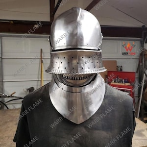 Puede incluir: Un casco medieval de acero pulido con visera y protector de cuello. El casco está diseñado para proteger la cabeza y el cuello. La visera tiene múltiples agujeros pequeños para ventilación. El casco se muestra sobre una camiseta negra.
