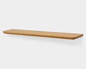 Könnte beinhalten: Ein schwebendes Holzregal mit natürlicher Holzmaserung. Das Regal ist rechteckig mit abgerundeten Kanten und an einer weißen Wand befestigt. Das Regal ist etwa 122 cm lang.