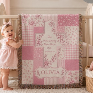 Könnte beinhalten: Eine rosa-weiß karierte Babydecke mit Blumen- und Vichy-Mustern. Die Decke trägt den Text "You shine among them like a star" und den Namen "Olivia". Die Decke wird in einem Kinderbett gezeigt.