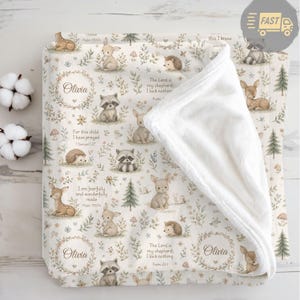 May include: A cream-coloured baby blanket with a woodland animal pattern, including deer, rabbits, and raccoons. The blanket features floral accents and text that reads "Olivia" and Bible verses. The reverse side is a soft, white material.