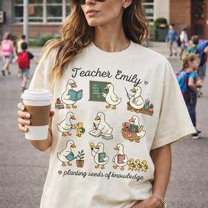 Könnte beinhalten: Cremefarbenes T-Shirt mit einem skurrilen Design mit Cartoon-Enten in verschiedenen Lehrerrollen. Das Shirt enthält den Text "Teacher Emily" und "planting seeds of knowledge."