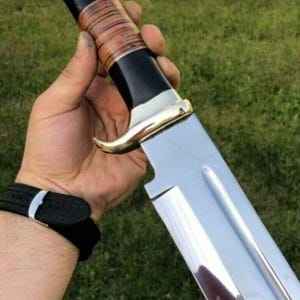 Könnte beinhalten: Ein großes, poliertes Stahlmesser mit schwarzem Griff, Akzenten aus goldfarbenem Metall und einem braunen, strukturierten Griff. Die Klinge ist breit und hat eine einzigartige Kerbe in der Nähe des Griffs. Das Messer wird vor grünem Gras gehalten.