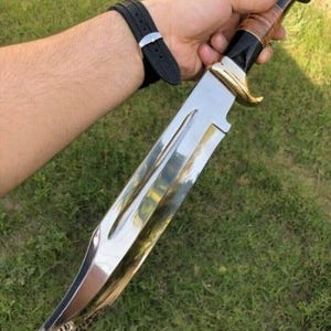 Pode incluir: Uma faca grande e prateada polida com uma pega castanha e preta e detalhes dourados. A lâmina é larga e curva, com um padrão decorativo perto da ponta. A faca é segurada contra um fundo de relva verde.