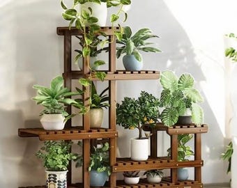 May include: A multi-tiered wooden plant stand displaying various potted plants. The stand is constructed from dark brown wood and features multiple shelves of varying sizes. The plants include diverse green foliage and flowering plants in white, blue, and patterned pots. The stand is on wheels.
