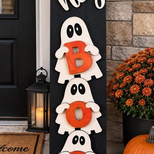Puede incluir: Un letrero de madera negro, alto y rectangular, con la palabra "hey" en letra cursiva blanca en la parte superior. Debajo, tres figuras de fantasmas blancos están apiladas verticalmente, cada una sosteniendo una letra naranja para formar "BOO".