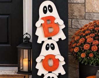 May include: A tall, rectangular black wooden sign featuring the white script word "hey" at the top. Below, three white ghost cutouts are stacked vertically, each holding a bright orange letter to spell out the word "BOO" in a column.