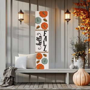 Puede incluir: Un letrero de porche alto y rectangular de madera blanca con el texto negro "hello FALL" en el centro. El letrero está decorado con calabazas en relieve pintadas en naranja y verde salvia, junto con pequeñas hojas marrones dispuestas alrededor.