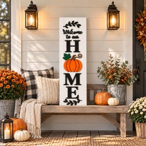 May include: A tall, rectangular white wooden sign with black text that reads Welcome to our HOME, featuring a bright orange pumpkin with a green leaf replacing the letter O. The sign is displayed on a wooden bench with pillows and pumpkins.