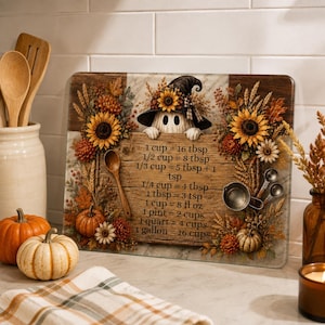 May include: A rectangular glass cutting board featuring a ghost wearing a witch hat, surrounded by sunflowers and autumn foliage. The center displays a wood-grain panel with kitchen conversion measurements including 1 cup equals 16 tbsp.