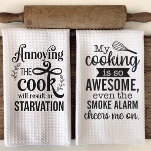 May include: Two white kitchen towels with black text. One towel reads "Annoying the cook will result in starvation." The other says "My cooking is so awesome, even the smoke alarm cheers me on." A wooden rolling pin rests above.
