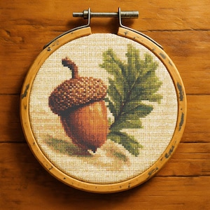 Puede incluir: Bordado de punto de cruz de una bellota y una hoja de roble, exhibido en un aro de madera redondo. La bellota es marrón y naranja, con una tapa texturizada. La hoja de roble es verde. El fondo es de tela beige claro.