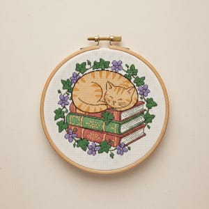 Peut inclure: Broderie au point de croix dans un cercle en bois représentant un chat tigré orange endormi sur une pile de livres. Les livres sont verts et rouges, entourés d'une couronne de feuilles vertes et de fleurs violettes. Le cercle mesure environ 15 cm de diamètre.