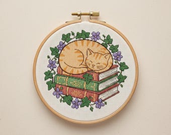 May include: Cross-stitch embroidery in a wooden hoop features a sleeping orange tabby cat atop a stack of books. The books are green and red, surrounded by a wreath of green leaves and purple flowers. The hoop is approximately 6 inches in diameter.