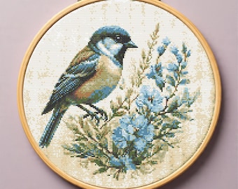 May include: A cross-stitch embroidery of a bird with blue, brown, and beige tones, perched on a branch with blue flowers. The artwork is set within a round wooden embroidery hoop, creating a framed piece.