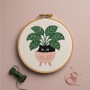 May include: A cross-stitch embroidery hoop showing a black cat in a pink plant pot with green leaves. The embroidery is set in a wooden hoop. A skein of green thread and a pink thread are on the pink background.