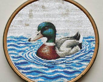 Könnte beinhalten: Kreuzstich-Stickerei einer Stockente, die in blauem Wasser schwimmt, in einem runden Holzreifen. Die Ente hat grüne, braune und weiße Details. Das Wasser hat ein Wellenmuster.