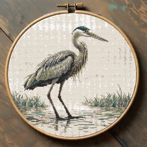 Könnte beinhalten: Kreuzstich-Stickerei eines Graureihers in einem Holzreifen. Der Reiher ist in Grau-, Blau- und Grüntönen mit detaillierten Stichen dargestellt. Der Hintergrund ist hellbeige, mit grünem Gras am Wasser. Der Reifen hat einen Durchmesser von ca. 20 cm.