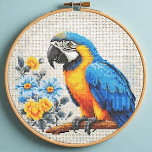 Puede incluir: Bordado de punto de cruz de un guacamayo azul y dorado posado en una rama, rodeado de flores azules y amarillas. La obra de arte está enmarcada en un aro de madera redondo, mostrando el detallado trabajo de aguja y los colores vibrantes.