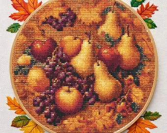 Puede incluir: Un bordado de punto de cruz en un aro de madera, que representa una naturaleza muerta de manzanas, peras y uvas. El diseño utiliza colores otoñales, con hojas naranjas, rojas y amarillas que rodean el arreglo de frutas. El bordado está sobre un fondo blanco.