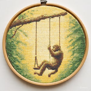 May include: A cross-stitch embroidery of a monkey swinging on a swing. The monkey is brown, and the background is yellow and green, with a wooden hoop frame.