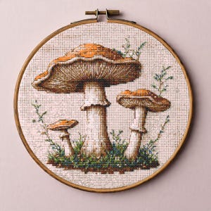 May include: Cross-stitch embroidery of three mushrooms in a wooden hoop. The mushrooms have brown and orange caps and white stems, set against a beige background with green foliage. The embroidery is framed in a round, wooden hoop.