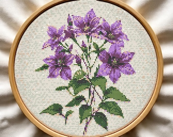 May include: A cross-stitch embroidery of purple flowers with green leaves and stems, set in a wooden embroidery hoop. The flowers are in full bloom, with detailed stitching creating a textured effect. The background is a light beige fabric.
