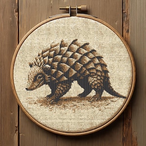 May include: A cross-stitch embroidery of a pangolin in shades of brown and black, set in a wooden hoop. The pangolin is depicted with detailed scales and is centered against a beige background, creating a rustic, handmade aesthetic.