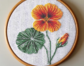 May include: Cross-stitch embroidery of a vibrant orange and red nasturtium flower, a green leaf, and a bud, set against a white background. The artwork is displayed in a round wooden embroidery hoop, showcasing a detailed floral design.