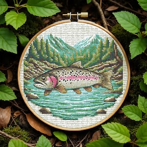 May include: A cross-stitch embroidery of a trout in a river scene, set in a wooden hoop. The fish has pink and white markings. The background features a forest and mountains, with shades of green, blue, and brown.