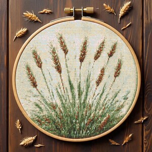 May include: A cross-stitch embroidery of wheat stalks in a wooden hoop. The wheat is depicted in shades of brown, green, and beige, set against a light background. The hoop is made of light-colored wood, and several wheat sprigs are scattered around it.