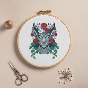 May include: A cross-stitch embroidery in a wooden hoop features a gray demon face adorned with red roses, white flowers, and green vines. The design includes horns and a stern expression. A thimble, scissors, and pins are arranged around the hoop.