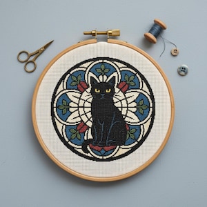 May include: A cross-stitch embroidery in a wooden hoop features a black cat with yellow eyes, centered within a stained-glass-style design of blue, white, and red floral elements. A pair of small gold scissors, a spool of blue thread, and buttons are placed around the hoop.