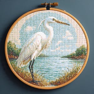 May include: A cross-stitch embroidery of a white egret standing near a body of water. The bird has a long beak and stands on black legs. The background features a blue sky with clouds and green foliage. The embroidery is set in a round wooden hoop.