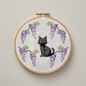 Puede incluir: Un bastidor de bordado de punto de cruz con un gato negro sentado debajo de flores de glicinas moradas colgantes. El gato tiene ojos amarillos y está centrado en un fondo de tela blanca. El bastidor es de madera.